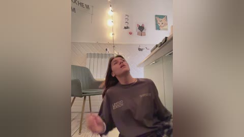 Media: Video of a young woman in a cozy living room, wearing a gray sweatshirt, leaning back with eyes closed, surrounded by string lights and a light blue chair.