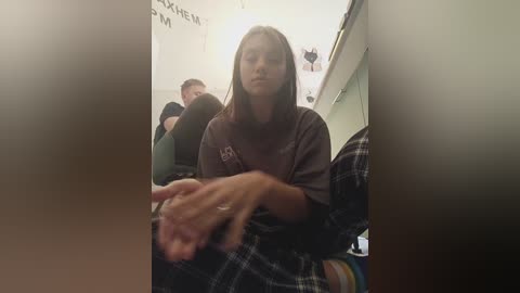 Media: Video of a young woman with straight, shoulder-length brown hair, wearing a brown t-shirt, sitting in a classroom, hands clasped, partially obscured by a blurry figure.