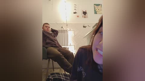 Media: Video of a man in a brown sweater and jeans, sitting on a stool in a dimly lit room with white walls, a table, and a partially visible woman in a checkered shirt.