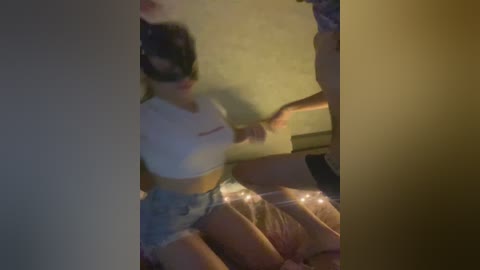Video of a young woman with fair skin, wearing a black mask, white crop top, and denim shorts, kneeling on a bed with a man's legs visible, dimly lit room.