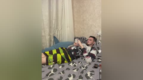 Media: Video of a young man lying in bed, wearing a patterned shirt and green and black striped pajama bottoms, playing a video game.