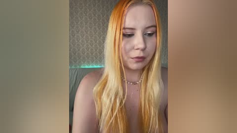 Video of a young Caucasian woman with long, straight, orange-blond hair, wearing a necklace, and a light green background with subtle, textured wallpaper. She has a neutral expression.