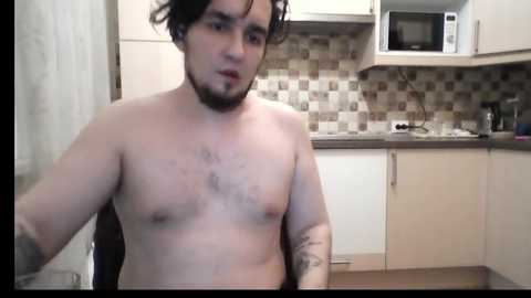 Video of a shirtless, light-skinned man with short, curly dark hair and a beard, in a modern kitchen with white cabinets, a microwave, and mosaic backsplash.
