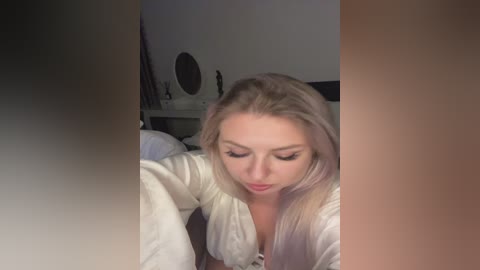 Media: A video of a young, fair-skinned woman with long, straight blonde hair, wearing a white, long-sleeved shirt, focusing intently on a tablet in a dimly lit bedroom with minimalistic decor.