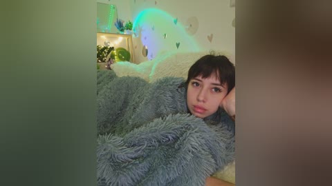 A young woman with short black hair, wearing a fluffy blue robe, is lying on a beige couch, surrounded by pastel-colored wall art and a lit lamp.
