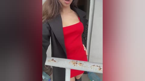 Media: Video of a woman with long brown hair, fair skin, wearing a tight red mini dress and a black blazer, standing on a balcony.