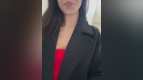 Media: Video of a woman with fair skin, dark hair, wearing a black blazer over a red top, partially obscured by a dark, blurred background.