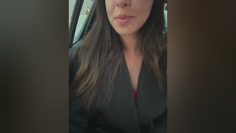 Media: Video of a woman with long brown hair, wearing a black blazer, sitting in a car. Her face is partially obscured, with a neutral expression. The background shows the car's interior.