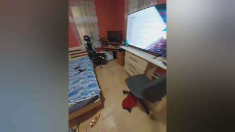 Video of a cluttered, small bedroom with a blue-patterned bed, a black office chair, a wooden desk with a computer monitor, and a large window with white curtains.