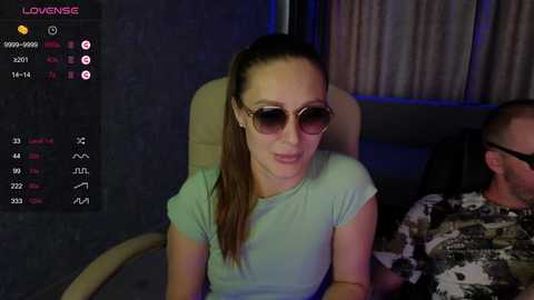 Video of a fair-skinned woman with long brown hair tied back, wearing aviator sunglasses and a light green t-shirt, seated indoors, looking slightly to the left, with a man in camouflage attire partially visible behind her.