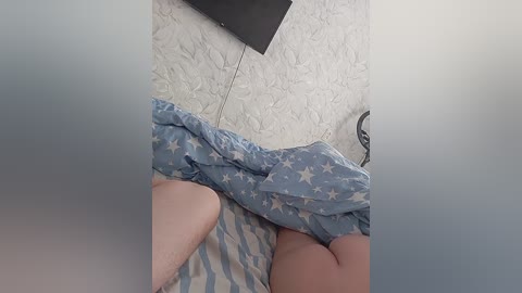 Video of a partially obscured person lying on a bed with blue and white star-patterned sheets. The person's legs are visible, and a wall-mounted flat-screen TV is in the background.