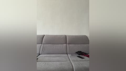Video of a minimalist, empty living room with a beige sectional sofa against a plain white wall. A black smartphone and a red object (possibly a piece of clothing) lie on the sofa's right side.