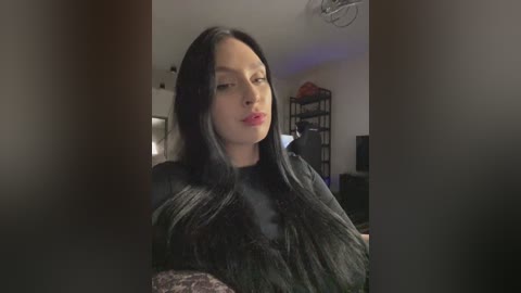 Video of a woman with long black hair, wearing a black dress, in a dimly lit living room with a black bookshelf and a red object on top.