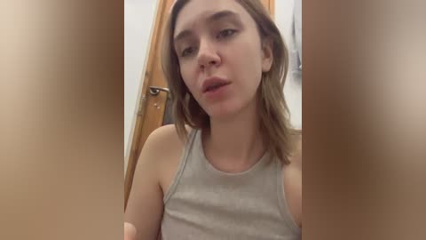 Media: A video of a young woman with fair skin and light brown hair, wearing a light gray tank top, standing in a bathroom with wooden doors and a towel rack in the background.