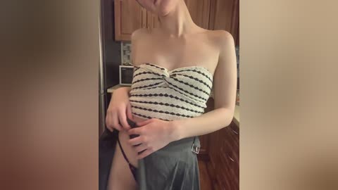 Media: Video of a slender, fair-skinned woman in a strapless, black-and-white striped top, pulling down her gray skirt to reveal black panties. Background shows a kitchen with wooden cabinets.