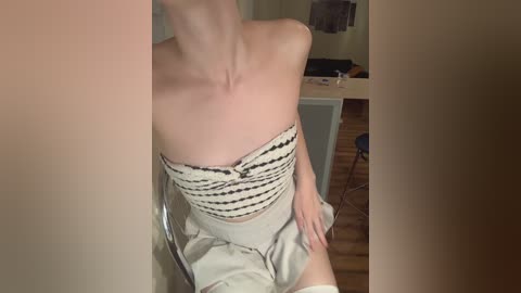 Media: Video of a slender woman with fair skin, wearing a strapless, beige top with black horizontal stripes and a high-waisted skirt, sitting on a transparent chair in a dimly lit room with wooden flooring.