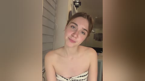 Media: Video of a young Caucasian woman with light skin and brown hair in a ponytail, wearing a strapless black and white patterned dress. She smiles softly in a dimly lit bathroom with white shutters and a towel rack in the background.