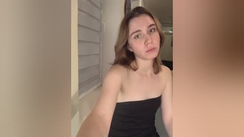 Media: A video of a young woman with shoulder-length brown hair, wearing a strapless black dress, sitting indoors next to a white shuttered window.