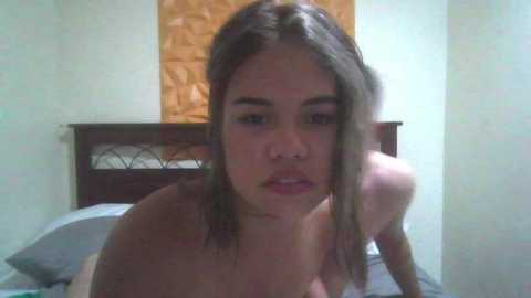 Video of a young woman with long brown hair, fair skin, and medium build, topless on a bed in a dimly lit room. The background features a wooden headboard and a patterned wall.