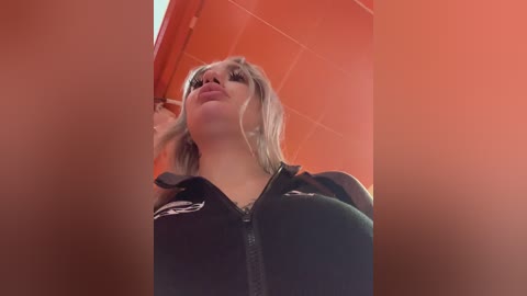 A video of a blonde woman in a black uniform, standing in a tiled room with an orange wall, looking upward. Her expression is neutral.