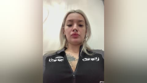 Video of a young woman with platinum blonde hair, wearing a black jacket with white tribal tattoos and a septum ring, set against a plain, light-colored background.