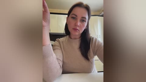 Media: Video of a fair-skinned woman with long brown hair, wearing a beige sweater, sitting at a table in a beige-walled room.