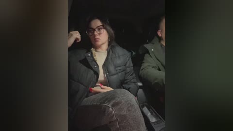 Media: Video of a woman in glasses, a puffy jacket, and high-waisted jeans, sitting in a dimly lit car. She looks contemplative, with a man in a green jacket beside her.