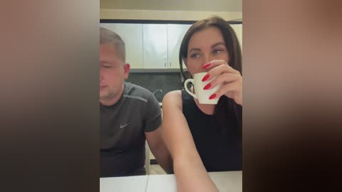 Media: A video of a man and woman in a kitchen. The man, with short hair, wears a gray t-shirt, eyes closed. The woman, with long brown hair, red nails, and a black top, drinks coffee.