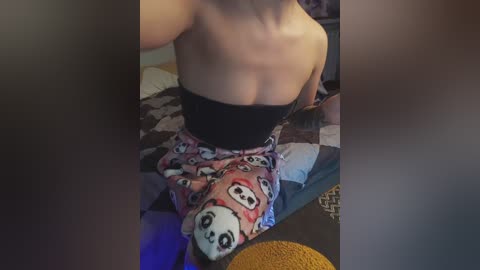 Video of a topless woman with fair skin, wearing black strapless bra and panda-themed pajama shorts, sitting on a bed with patterned sheets, in a dimly lit room.