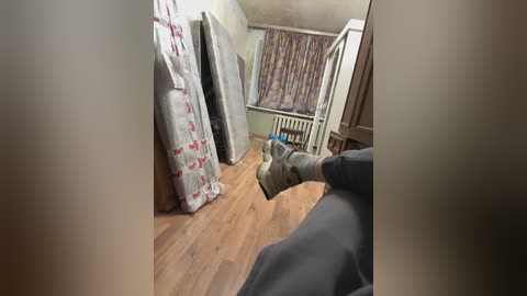 Media: A video of a cluttered, dimly lit room with a wooden floor. A person in dark clothing is sitting, their legs visible. Walls are plain, with a radiator and a curtain with red and white patterns.