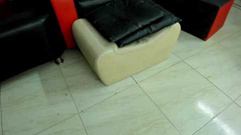 A video shows a beige, cushioned footrest with a black cushion on a tiled floor, part of a modern, minimalist indoor space with black and red furniture.