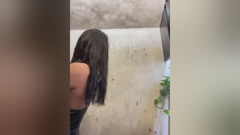 Media: Video of a woman with long black hair, wearing a black top, urinating in a public restroom with a concrete wall and a potted plant beside the urinal.