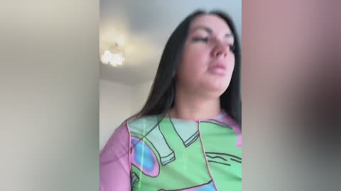 Video of a woman with long black hair, wearing a colorful abstract-patterned shirt, standing in a dimly lit room with a ceiling light.