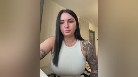 A video of a young woman with long black hair, wearing a white ribbed tank top, showcasing her large breasts and multiple tattoos on her arms. She stands in a narrow hallway with beige walls.