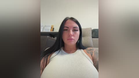 Video of a young woman with long black hair, fair skin, and a tattoo on her left arm, wearing a white ribbed tank top, sitting on a beige couch in a living room.