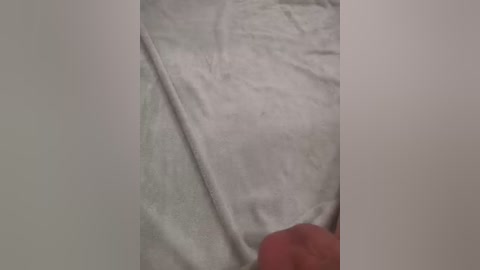 A video of a white, slightly crumpled fabric, possibly a bedsheet, with a diagonal crease in the center, partially revealing a faintly visible pattern. The background is plain and neutral, suggesting a simple, uncluttered setting.