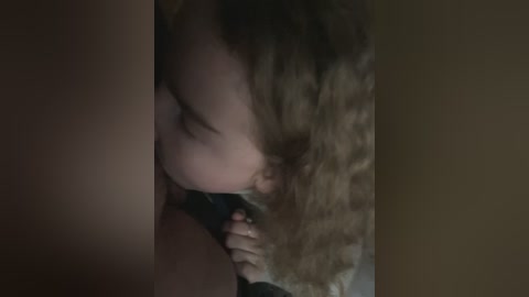 Media: Video of a young girl with curly, light brown hair, lying on a dark surface, eyes closed, possibly sleeping. The image is dimly lit, with a warm, muted color palette.