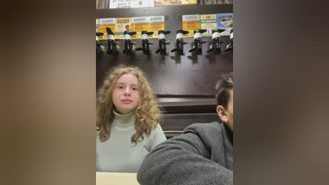 Media: Video of a young woman with curly blonde hair and fair skin, wearing a light gray turtleneck sweater, seated at a coffee counter. Background features a row of black coffee machines with yellow and blue labels.