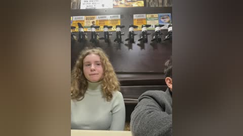 Media: Video of a young woman with curly blonde hair, wearing a beige turtleneck sweater, seated at a desk in a dimly lit room with a row of black microphones above her.