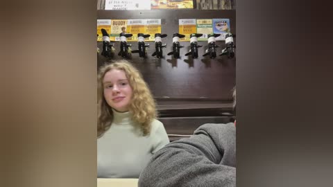 Media: Video of a Caucasian woman with curly blonde hair in a beige turtleneck sweater, seated in a fast-food restaurant with a soda fountain and menu boards in the background.