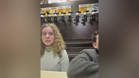 Media: A video shows a woman with curly hair in a gray sweater sitting next to a man with short hair in a gray shirt. Behind them, a dark wooden wall features a row of black figurines and yellow signs.