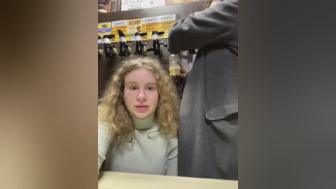 Media: Video of a young woman with curly blonde hair, wearing a beige turtleneck, seated in a pub. Behind her, a bartender in a gray shirt pours beer from a tap. The background includes beer posters and a counter.
