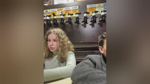 Media: Video of a young girl with curly blonde hair wearing a green sweater, seated at a table, with a man's arm visible, in a bar with beer taps and signs in the background.
