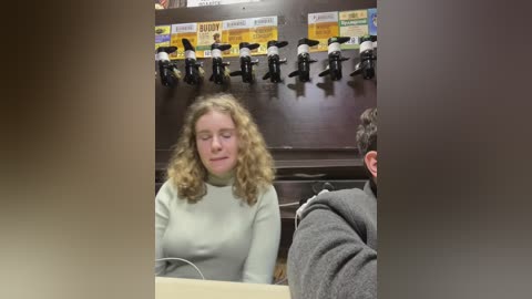 Media: Video of a woman with curly blonde hair and a light sweater, sitting in a dark bar with beer taps and a man partially visible.