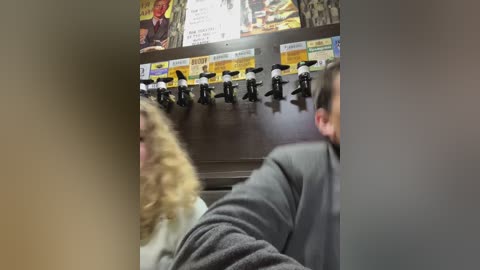 Media: Video of a man with short hair, wearing a gray shirt, leaning over a row of black and white soda dispensers in a dimly lit room.