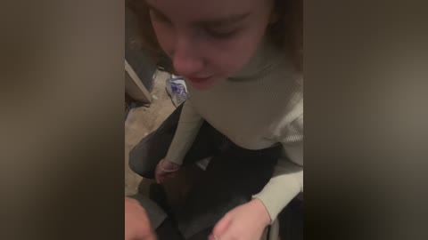Media: A video shows a young, fair-skinned girl with light brown hair in a beige sweater, seated on a wooden floor, holding a black stuffed animal. The background is dimly lit, suggesting a casual indoor setting.