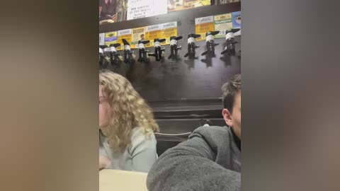 Media: Video of a classroom with a teacher wearing a white shirt, blonde student with curly hair, and a student in a gray hoodie. Background features a large screen displaying a video of students walking in a line.