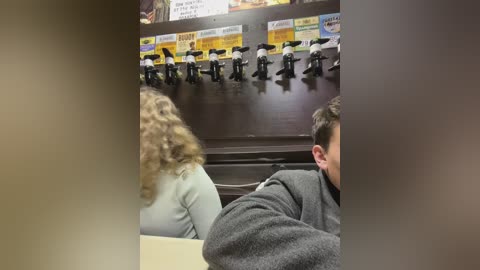 Media: Video of a dimly lit restaurant showing a waitress with curly hair in a light gray sweater, a man in a gray shirt, and a wall display of plastic toy soldiers with yellow and white signs.