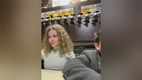 Media: Video of a young woman with curly blonde hair, wearing a white sweater, sitting at a counter in a diner. Behind her, a row of black beer taps is visible.