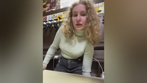 Media: Video of a young woman with curly blonde hair, wearing a light green turtleneck sweater and high-waisted jeans, sitting at a table in a dimly lit room with a TV and posters on the wall.